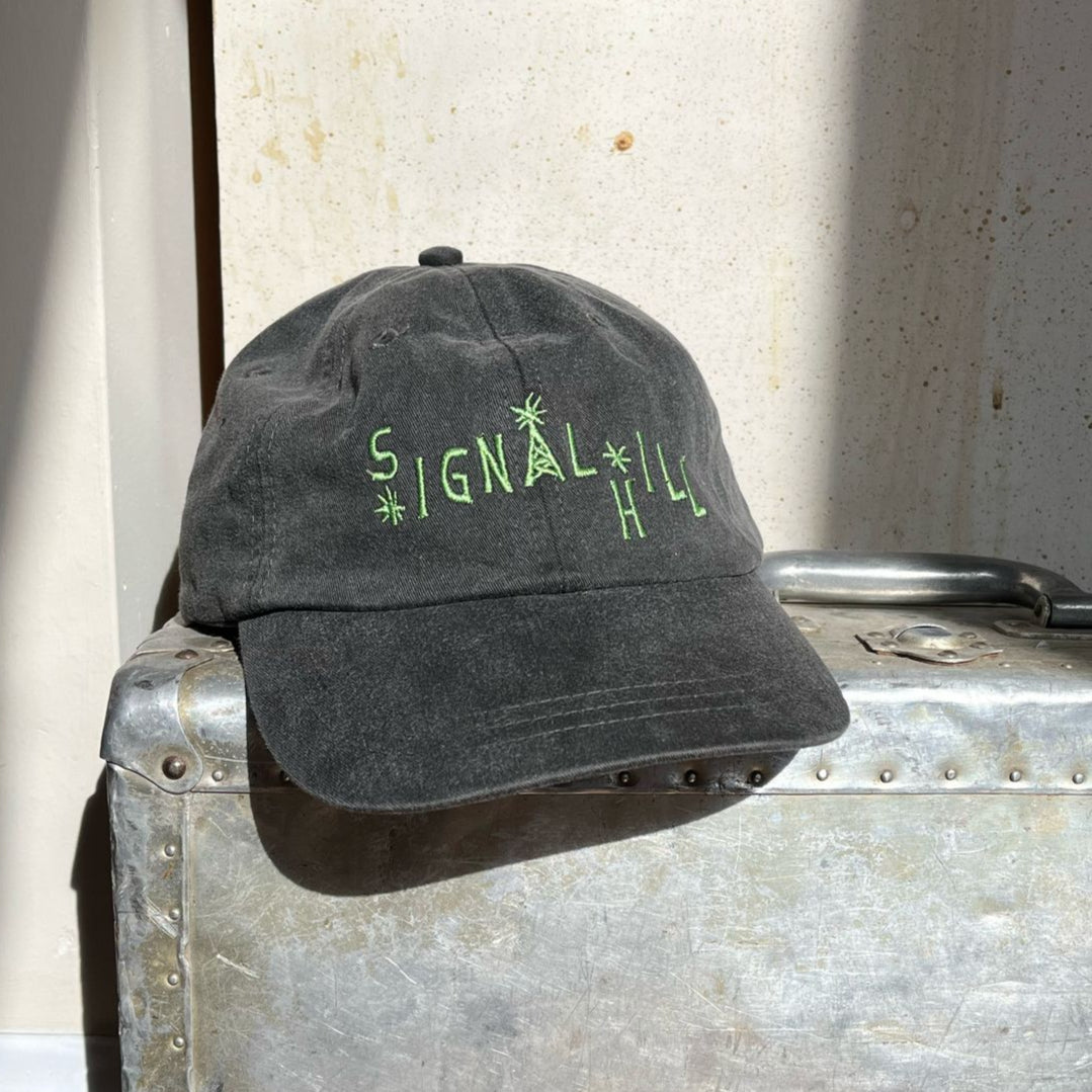 A dark grey baseball cap with Signal Hill written on it in green embroidery, sitting on a metal suitcase.